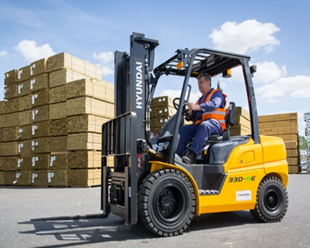 Operator in a hi-vis vest driving a Hyundai 33D-9 diesel counterbalance forklift past stacked timber
